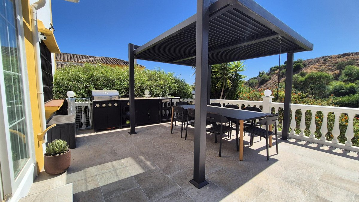 Modern outdoor dining area with BBQ station, covered by a pergola, overlooking a scenic hillside view.