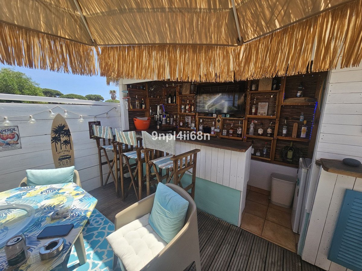 Outdoor bar with beach decor, surfboards, and seating area. Fully stocked shelves with bottles and TV screen display.