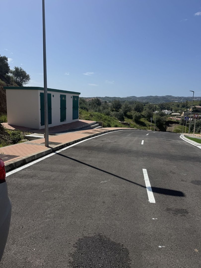 A small white building with green doors and a paved area in front, overlooking a hilly landscape under a clear blue sky.