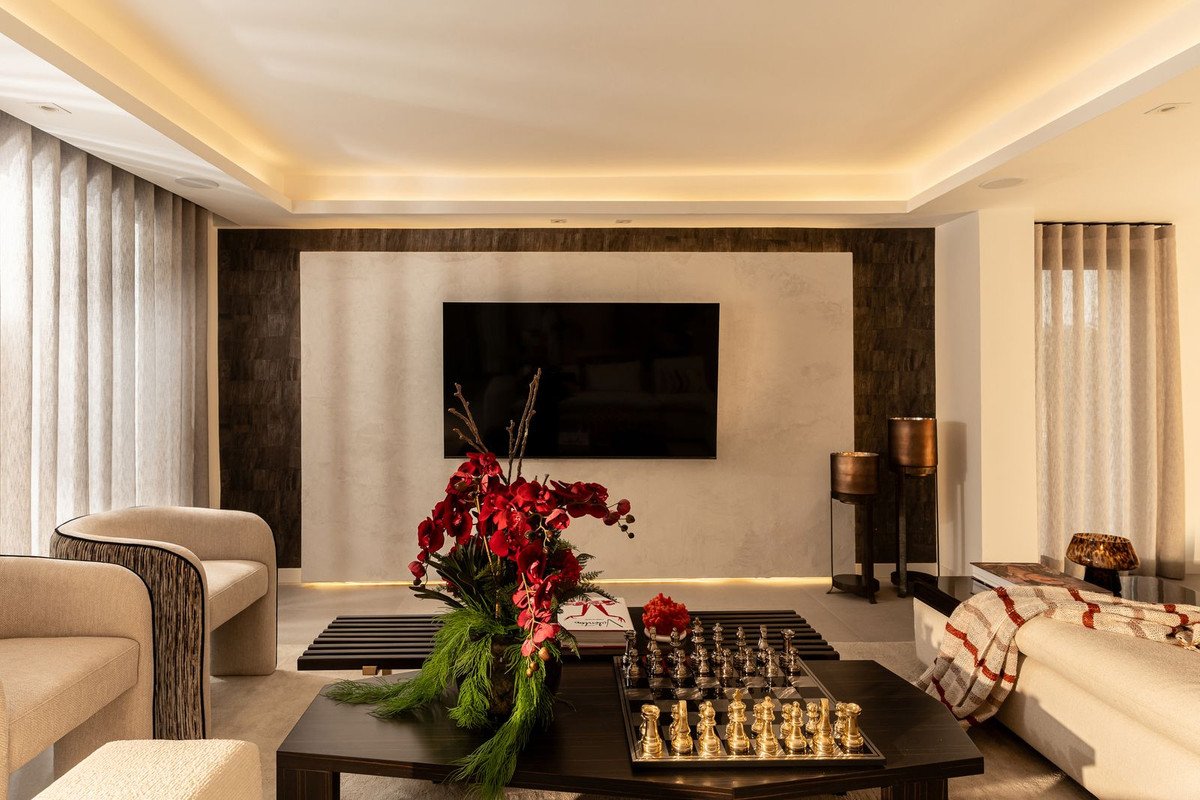 Modern living room featuring a large TV, luxurious armchairs, a coffee table with a chess set, and a vase of red orchids.
