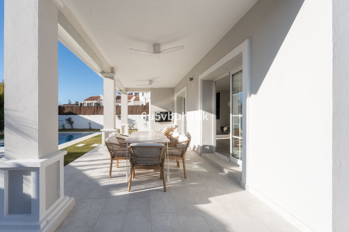 Covered terrace with dining table and chairs overlooking a swimming pool and garden on a sunny day.