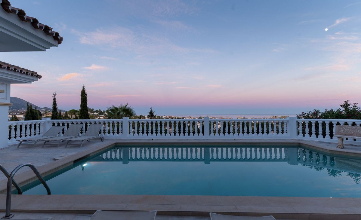 Swimming pool and terrace with lounge chairs overlooking the sea at sunset. Lush greenery and distant town visible.