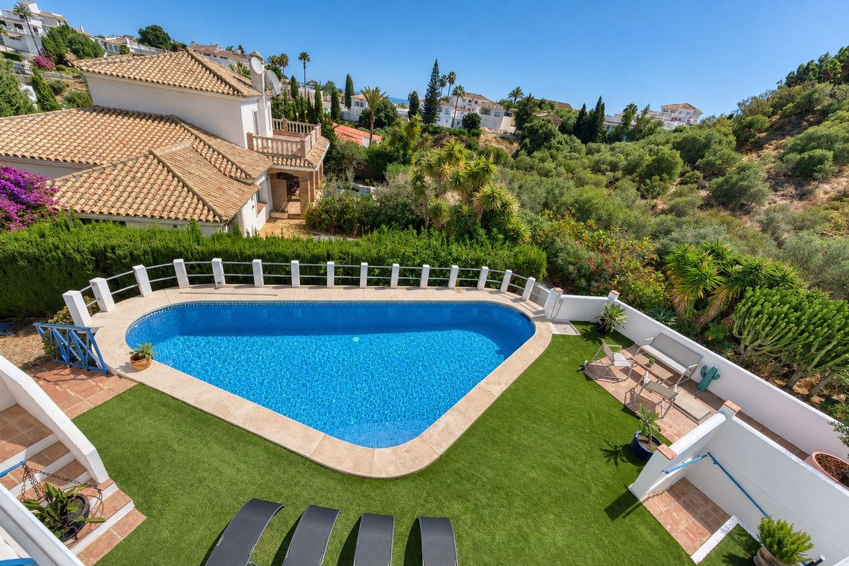A private swimming pool surrounded by artificial grass, loungers, and seating area, with a villa and lush greenery in the background.