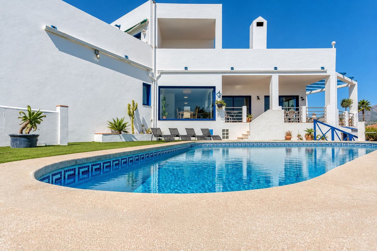 A white villa with blue window frames and a large swimming pool in front. Sun loungers are by the pool.