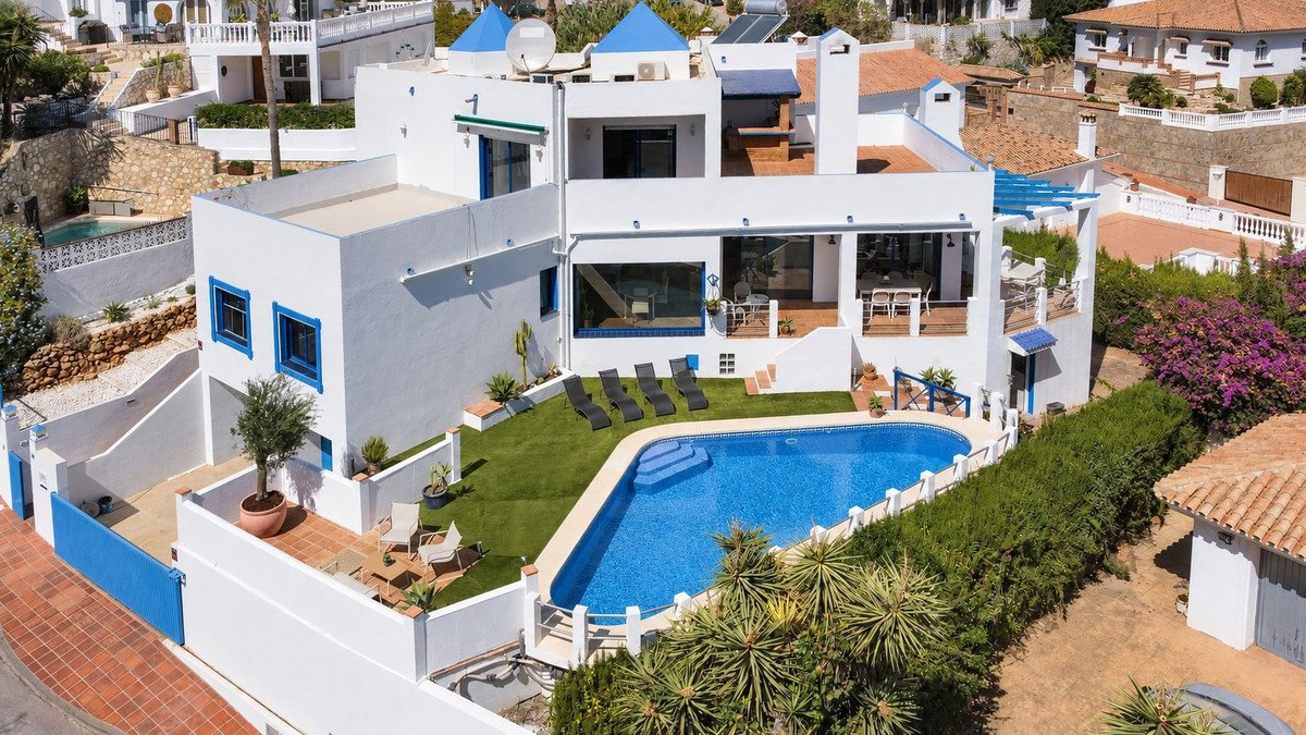 White villa with blue accents, featuring a swimming pool, sun loungers, outdoor dining area, and lush garden with sea views.