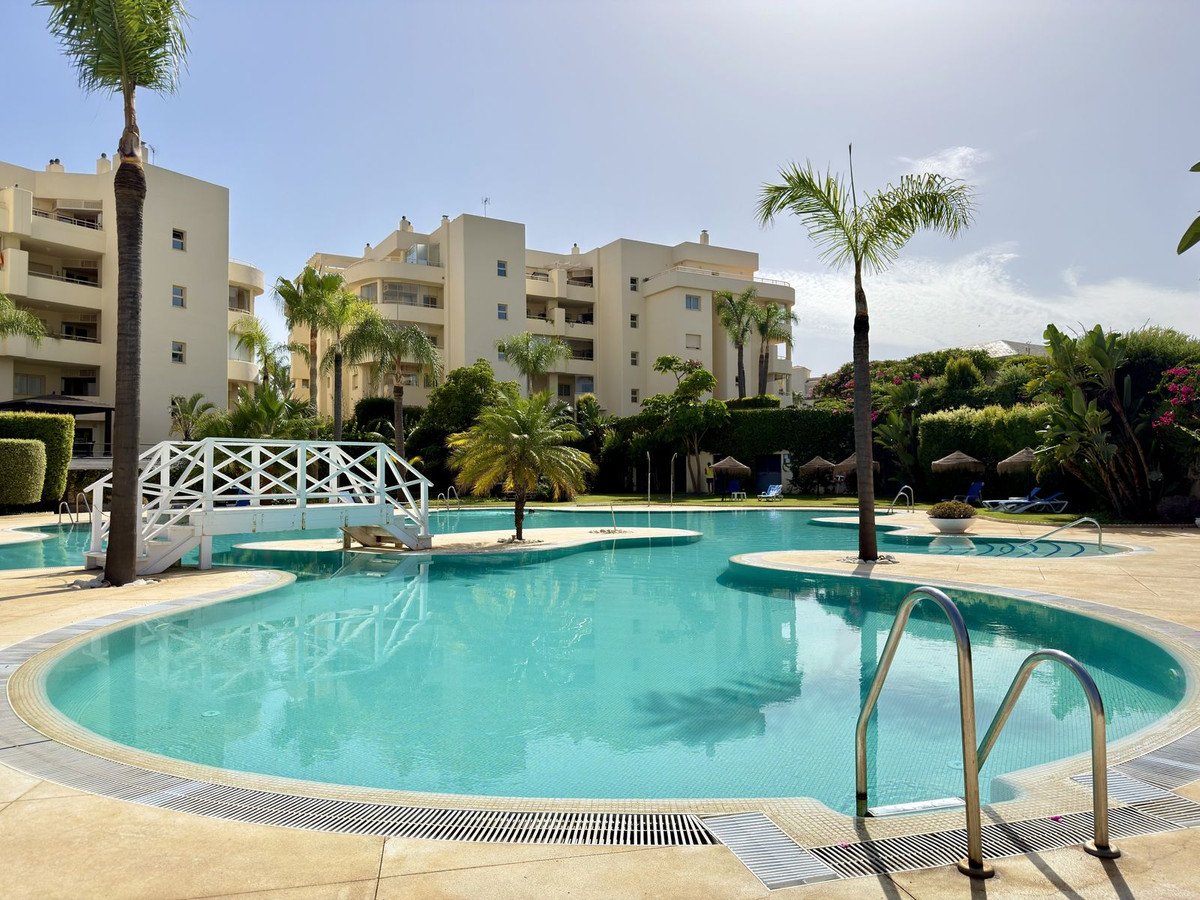 A resort complex with a large, sparkling blue swimming pool featuring a white decorative bridge and surrounded by palm trees and lush greenery.