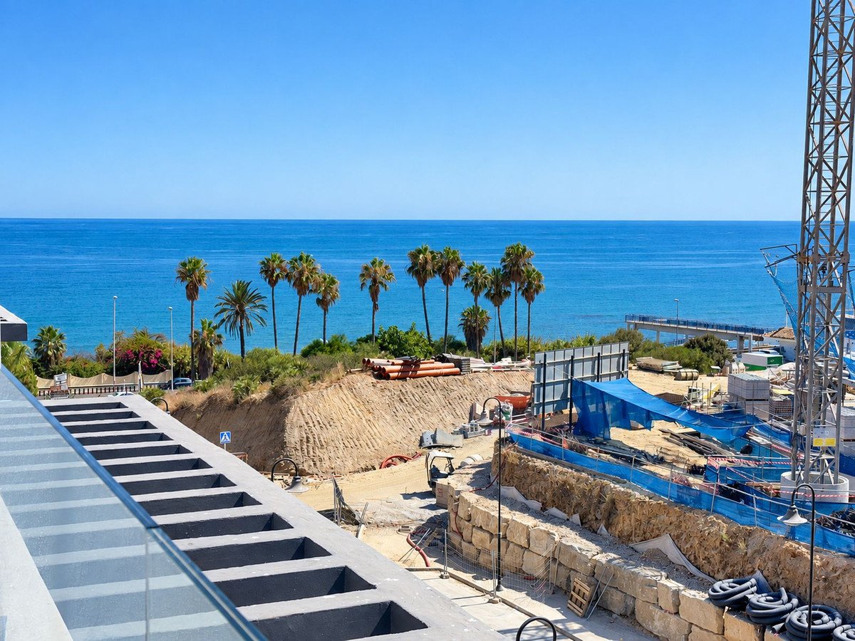 A balcony view overlooking the sea with palm trees, a construction site, and a clear blue sky.