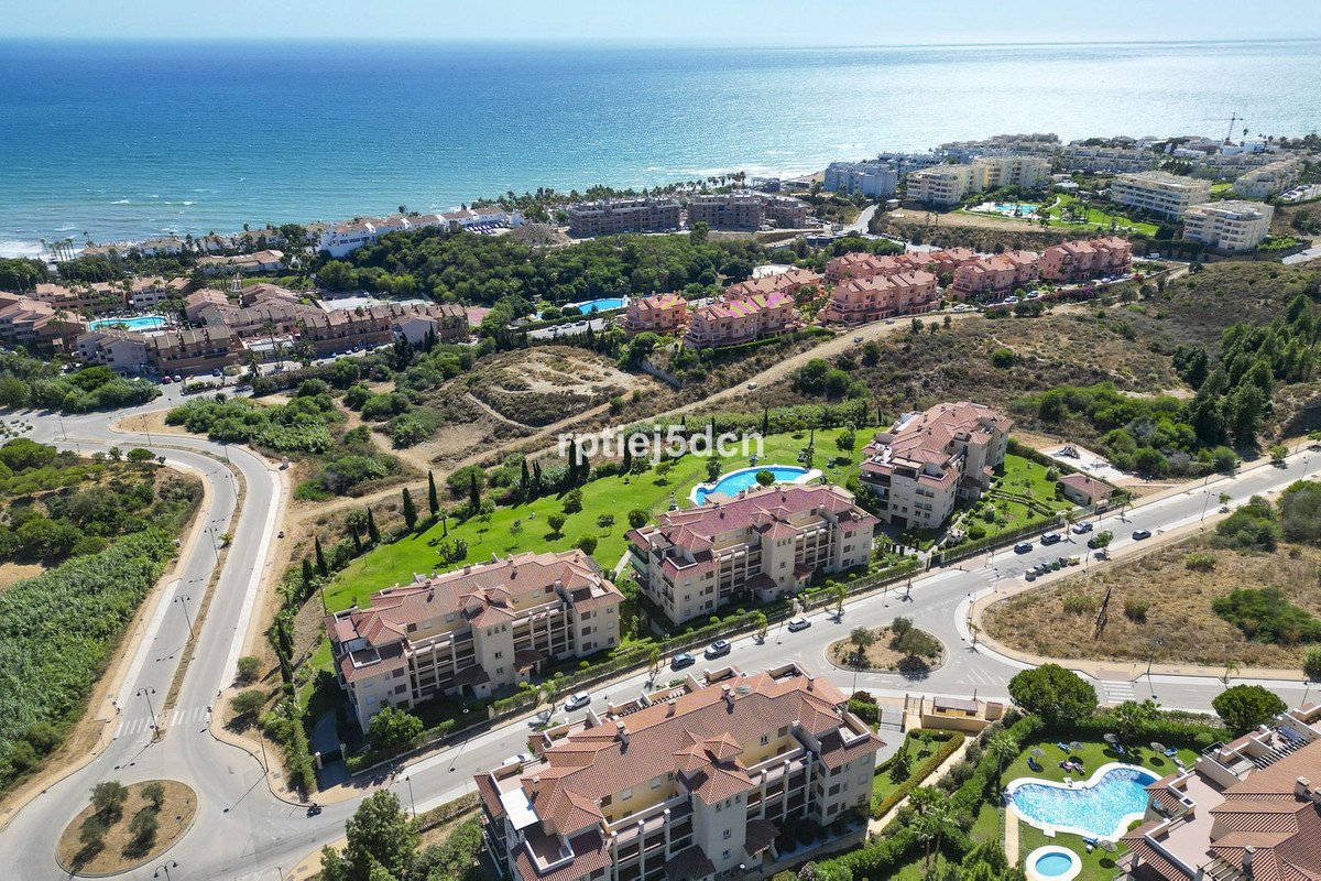 Aerial view of modern apartment buildings with swimming pools and lush green surroundings, overlooking the blue sea on a sunny day.