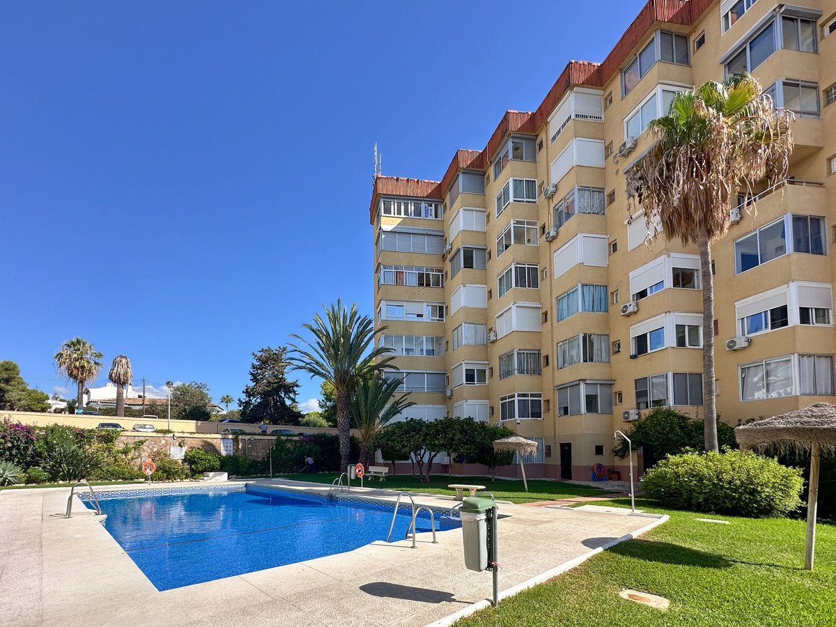 Exterior view of a yellow apartment building with balconies, overlooking a blue swimming pool and palm trees under a clear blue sky.