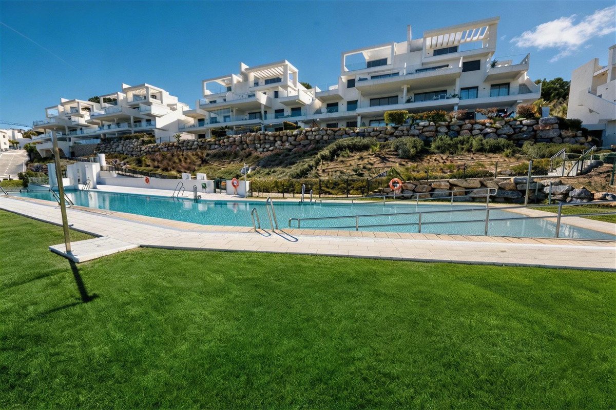 Modern white apartment buildings with balconies overlooking a swimming pool and manicured lawns under a clear blue sky.