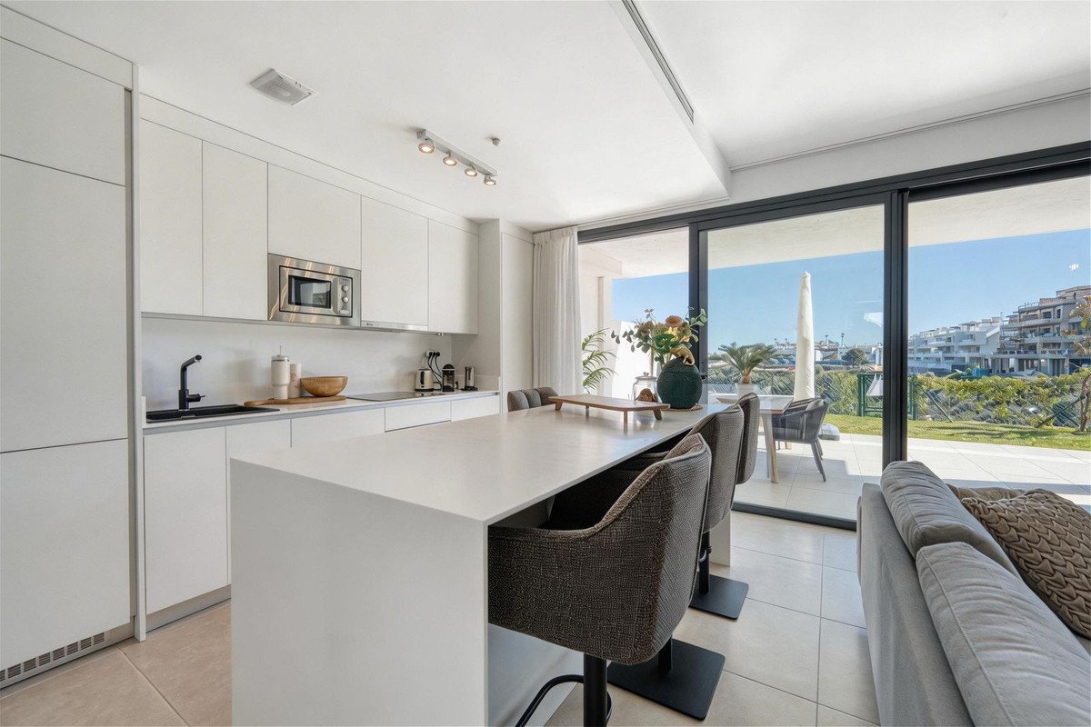 Modern white kitchen with island seating, overlooking a terrace and sea view with distant buildings.