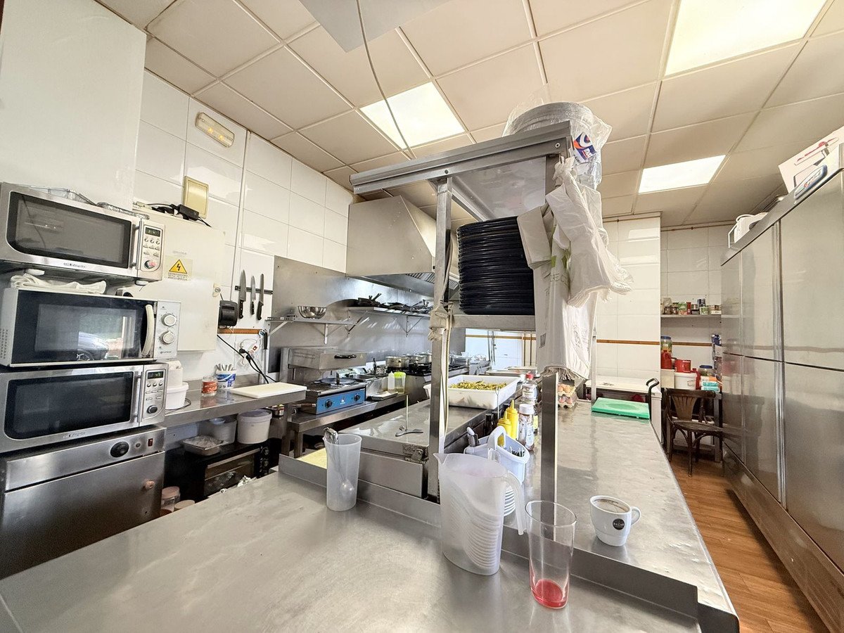 Commercial kitchen with stacked microwaves, stainless steel counters, gas stoves, and overhead shelving with utensils and supplies.