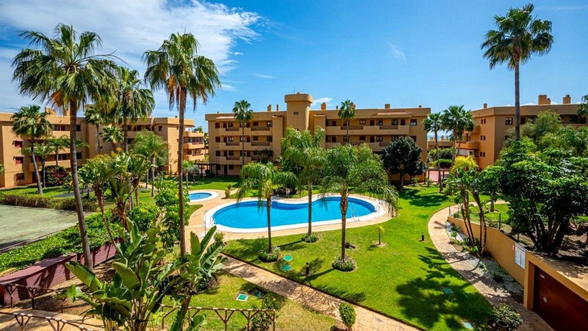 Apartment complex with palm trees surrounding a blue swimming pool and manicured lawns on a sunny day.