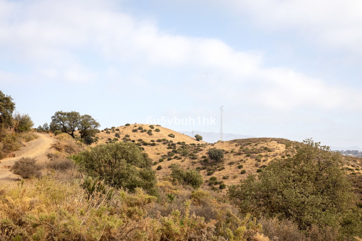 A dirt path winds through dry, rolling hills covered in scrub and trees under a partly cloudy sky.