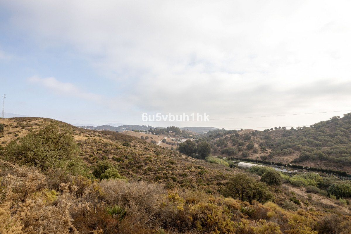 A wide rural landscape with rolling hills, scattered trees, and a cloudy sky overhead.