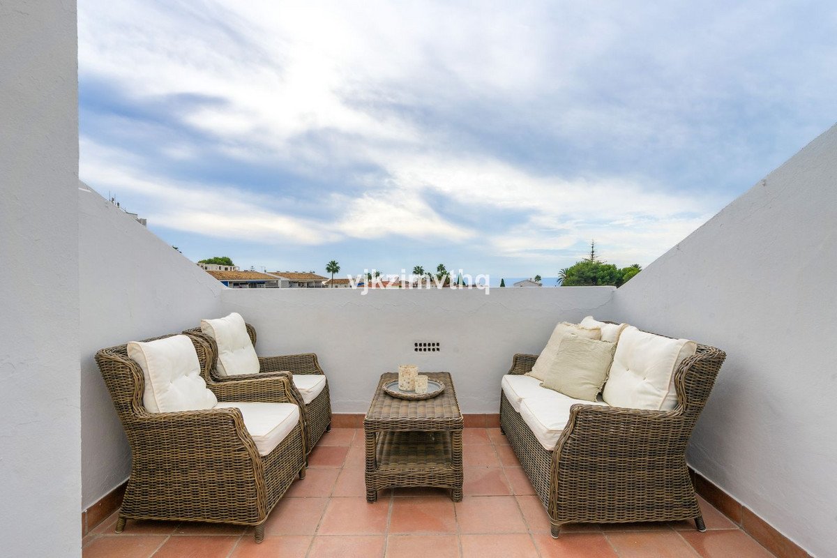 A terrace with wicker seating and a coffee table, offering a distant view of the sea and coastline under a cloudy sky.