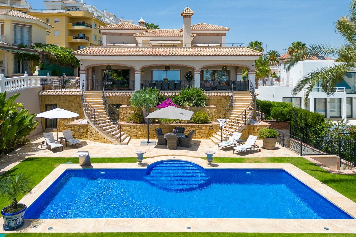 Luxury villa exterior with a large blue swimming pool, sun loungers, outdoor dining area, and manicured gardens under a clear sky.
