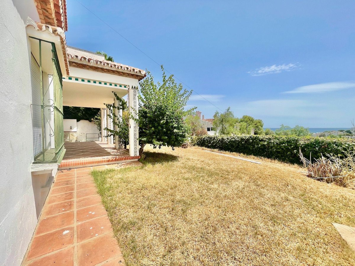 Exterior view of a covered terrace and a garden with a distant sea view under a clear blue sky.