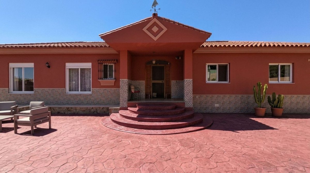 Exterior view of a house with a red terrace, seating area, cacti, and a wide entrance with steps under a clear blue sky.