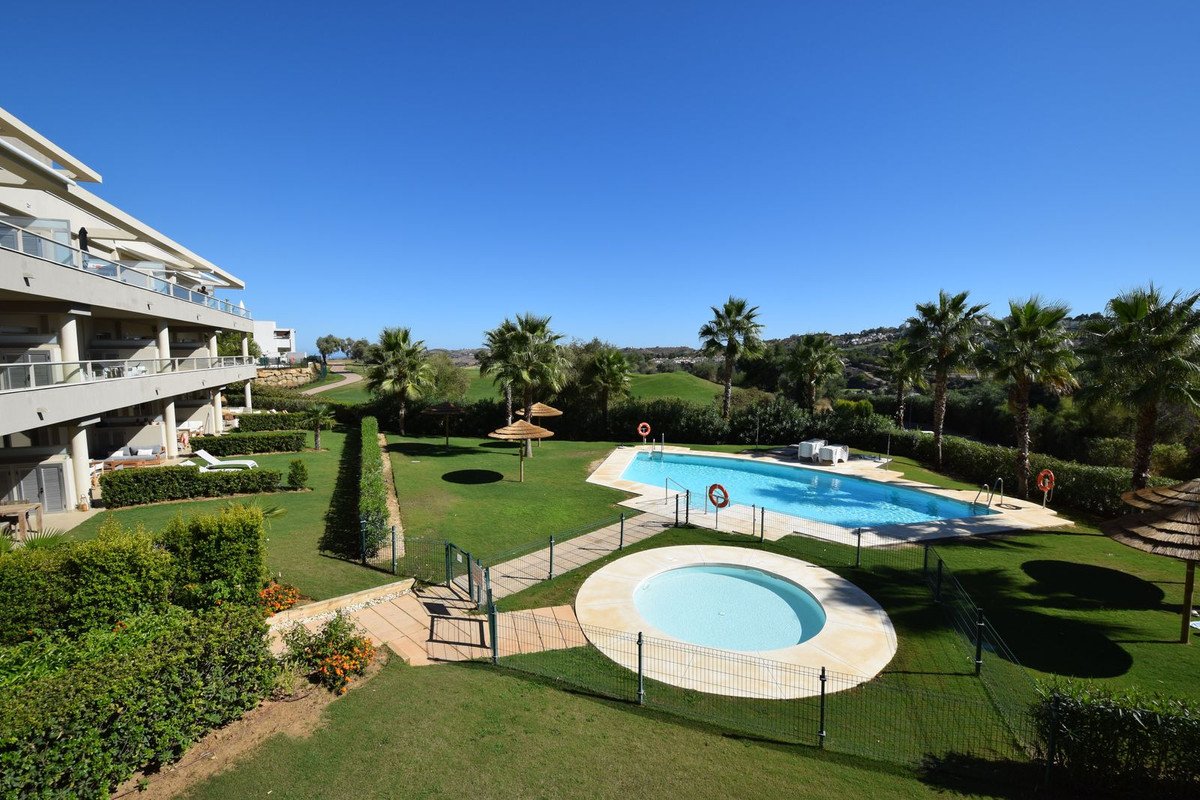Apartment building with balconies overlooking a swimming pool, small jacuzzi, and lush green gardens with palm trees and a golf course in the background.