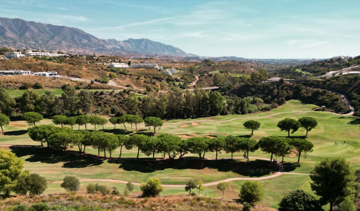 Lush green golf course with rolling fairways, scattered trees, and a backdrop of mountains under a clear blue sky.