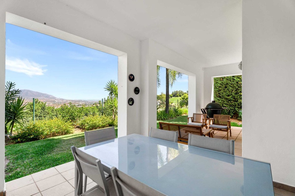 A covered terrace with a dining table and chairs, offering a view of mountains and greenery.