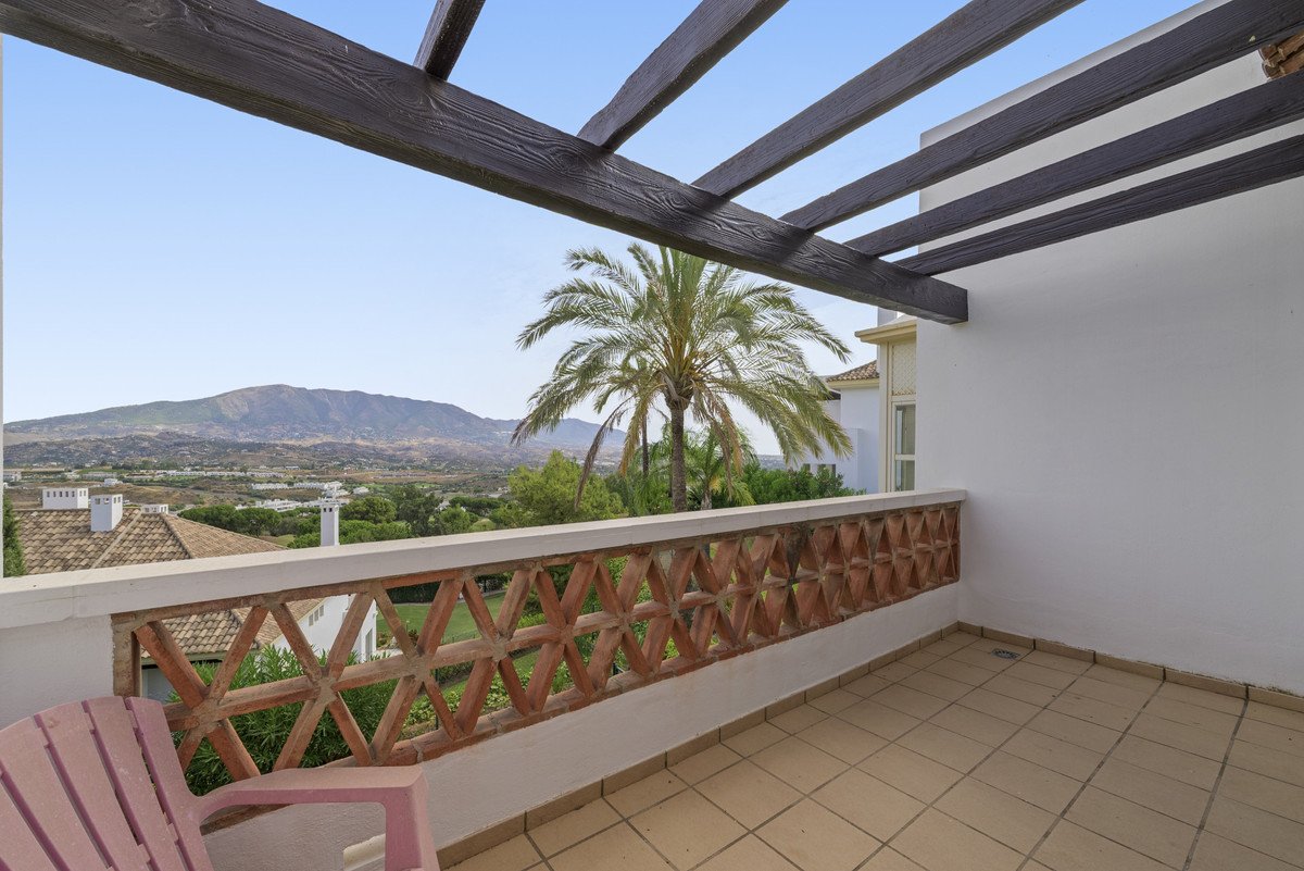 A covered balcony with a decorative railing offers a view of mountains, palm trees, and surrounding greenery.