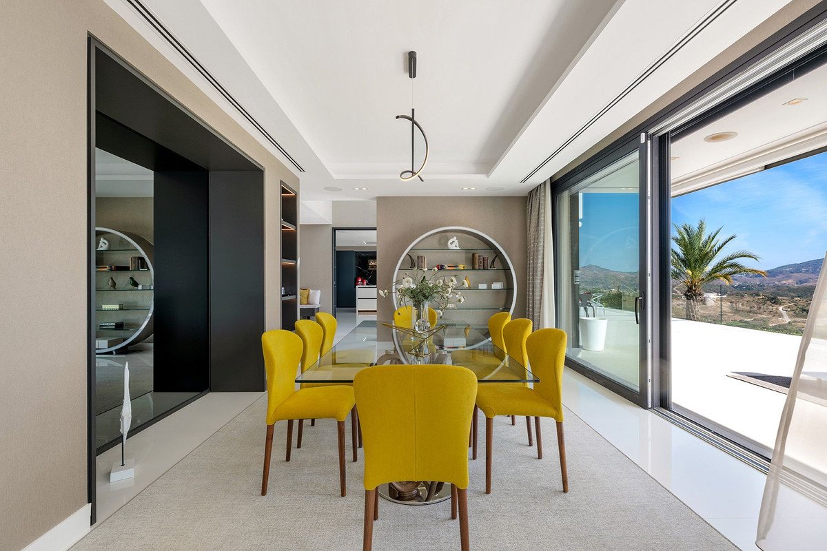 Modern dining room with yellow chairs, glass table, and a stunning view of the sea and palm trees through sliding glass doors.