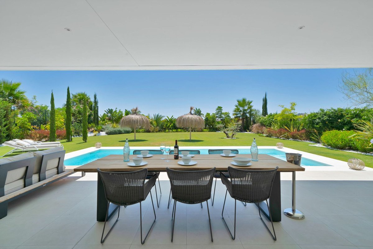 Outdoor dining area with table and chairs overlooking a swimming pool and garden on a sunny day.