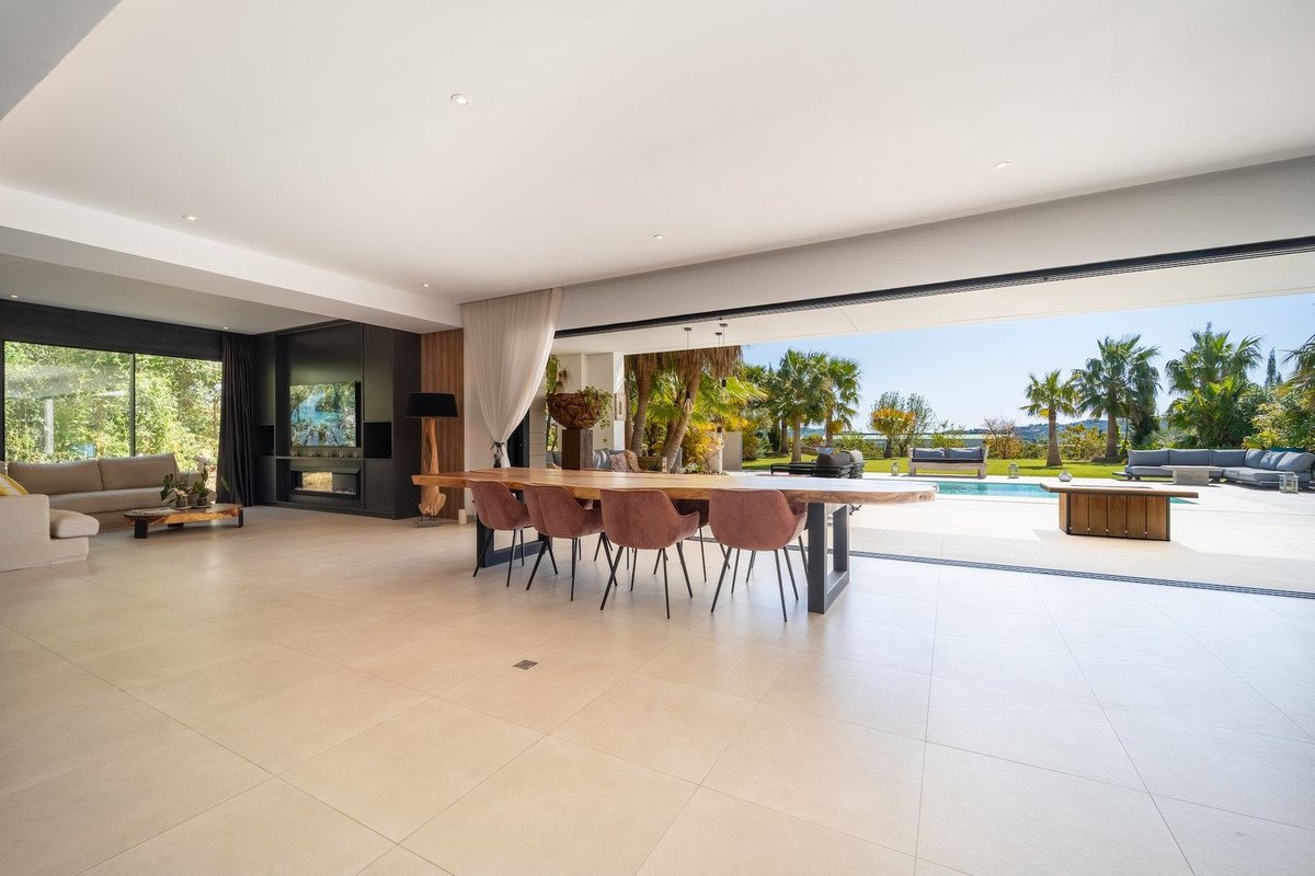 Spacious open-plan living and dining area with a large wooden table, opening onto a patio with a swimming pool and palm trees.