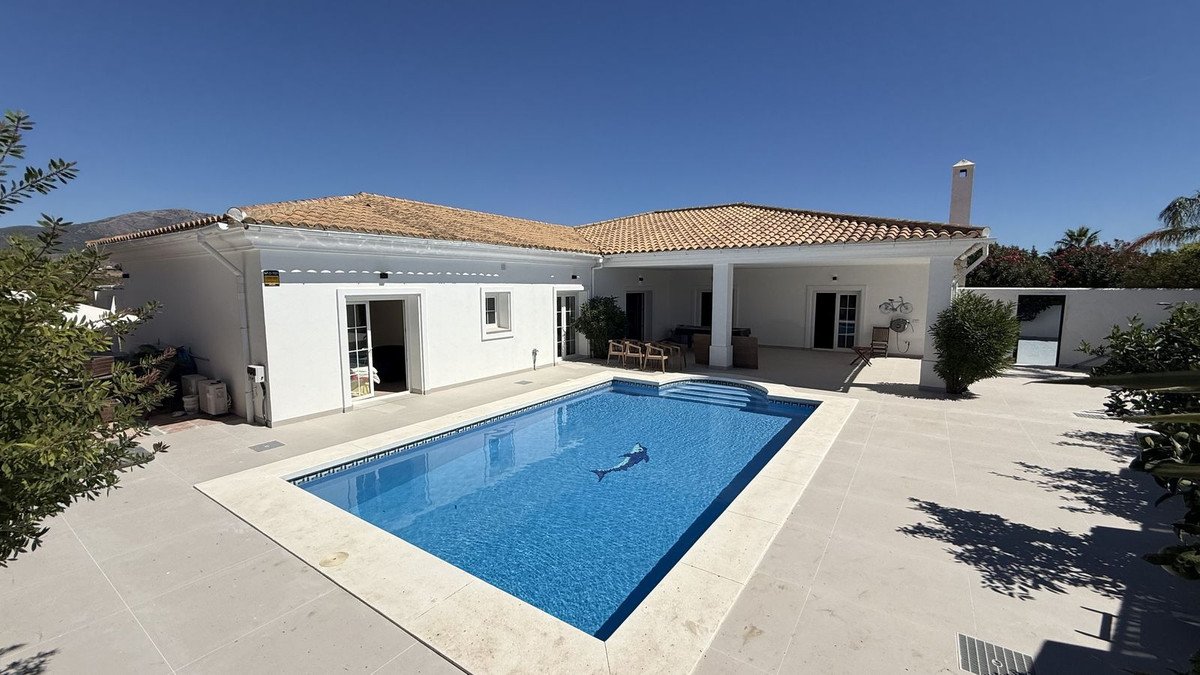 A white villa with a large blue swimming pool and a spacious tiled patio, surrounded by greenery under a clear blue sky.