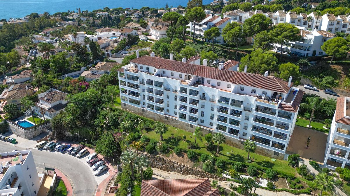 An aerial view of a white apartment building with multiple balconies and terraces, overlooking the sea and surrounded by greenery.