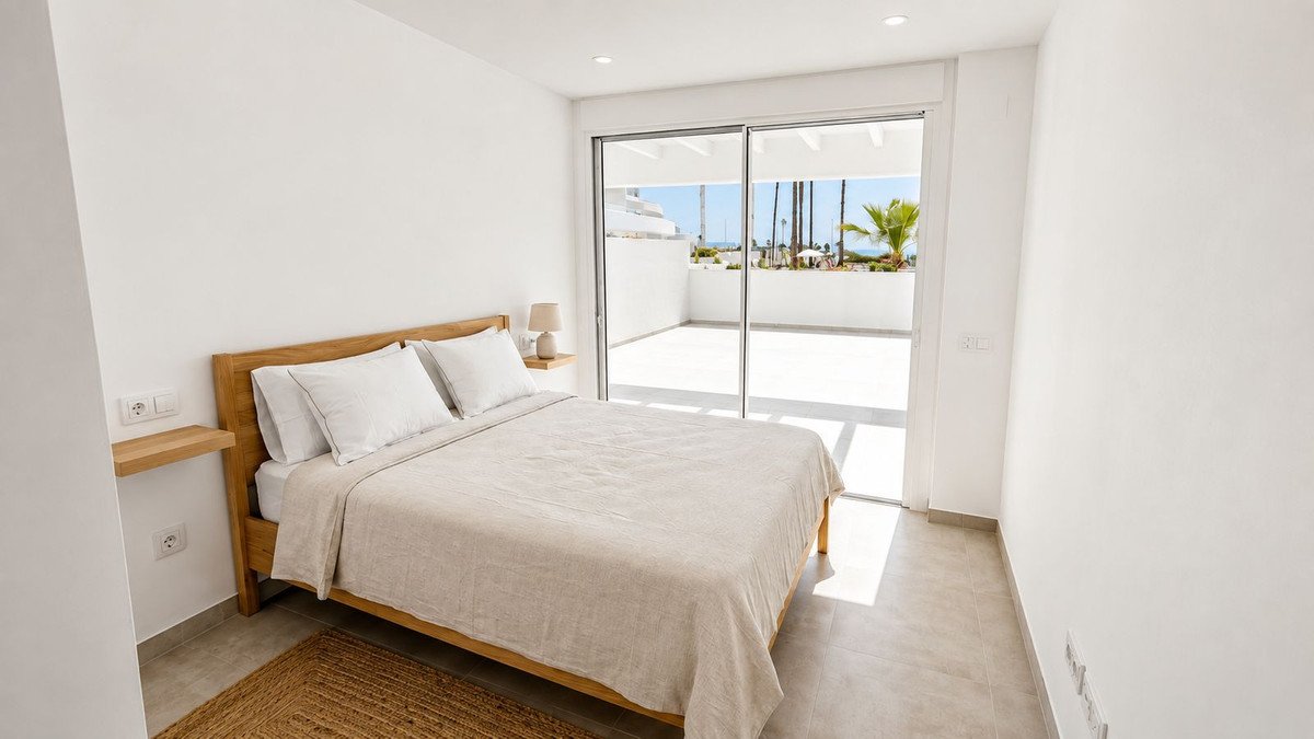 A minimalist bedroom with a queen-size bed, featuring a sliding glass door leading to a sunny terrace with palm trees and a sea view.