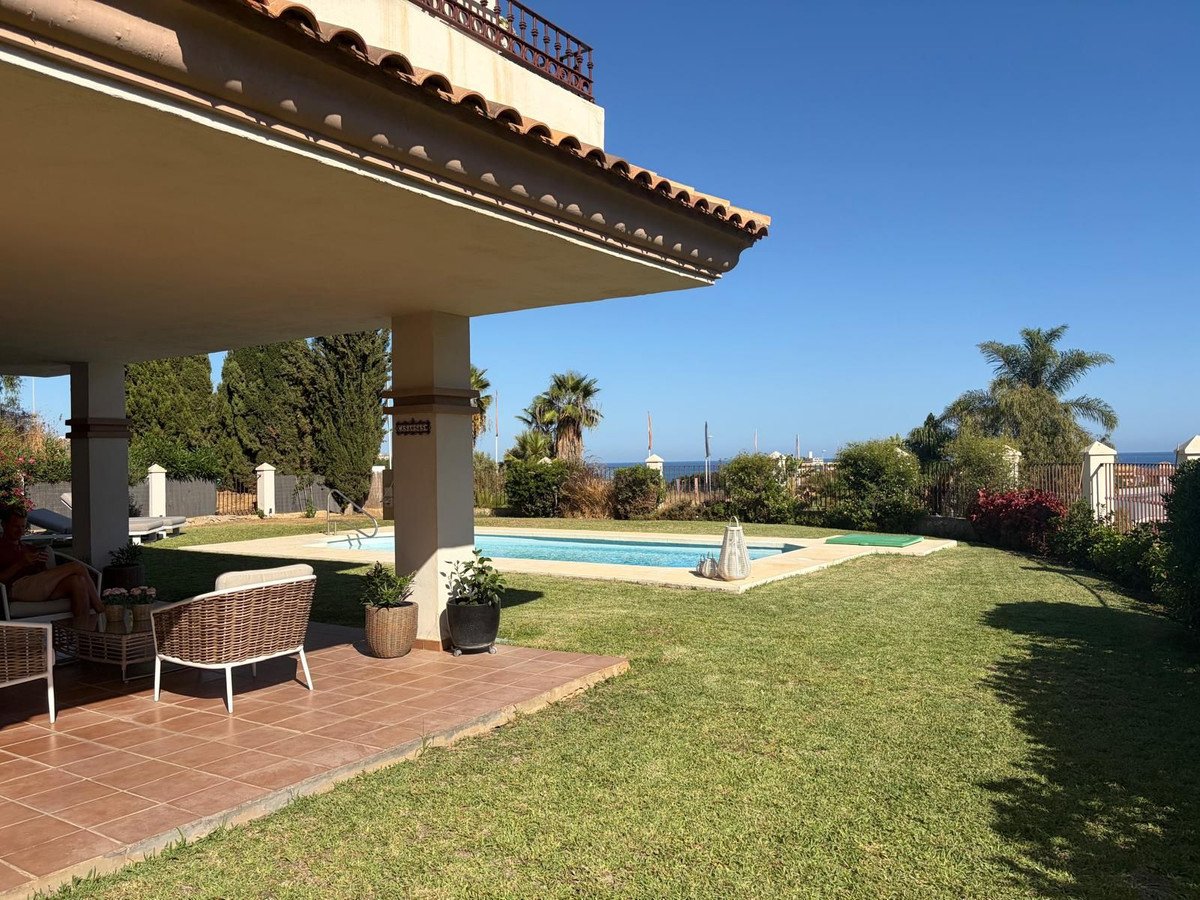 A covered patio with outdoor furniture overlooks a lush green lawn and swimming pool, with a distant sea view under a clear blue sky.