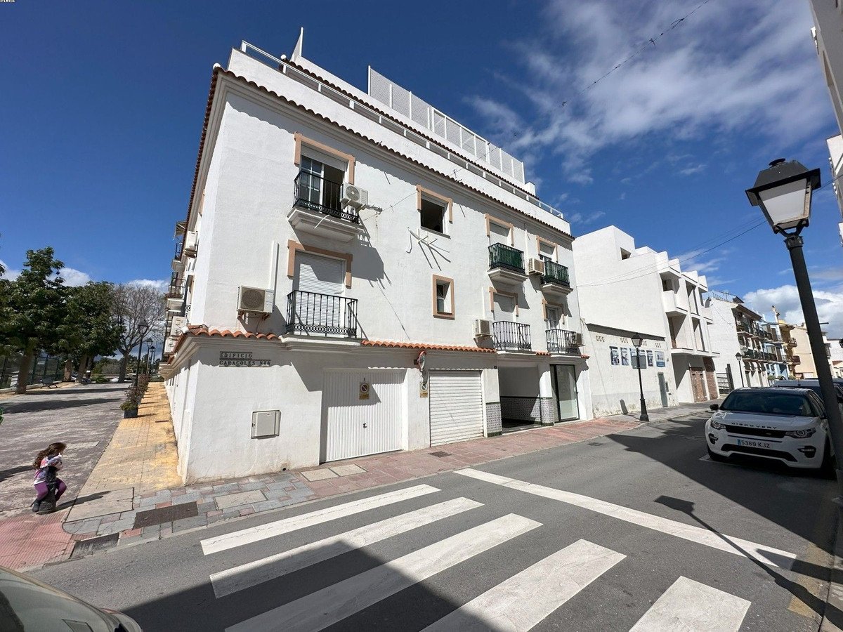 Exterior view of a white multi-story building on a street with balconies and air conditioning units, under a blue sky.