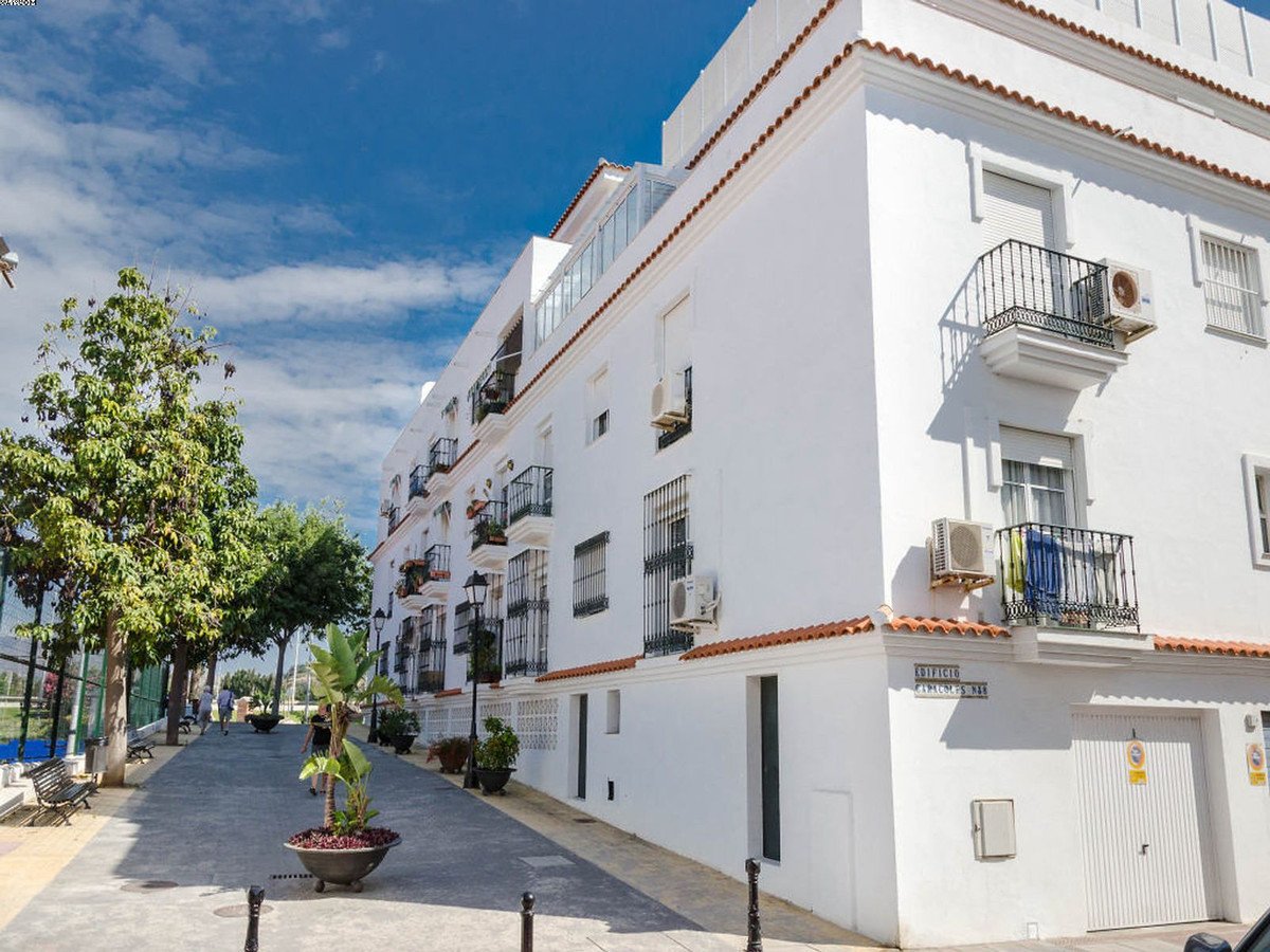 Exterior view of white apartments with balconies, air conditioning units, and a street with trees and people walking.