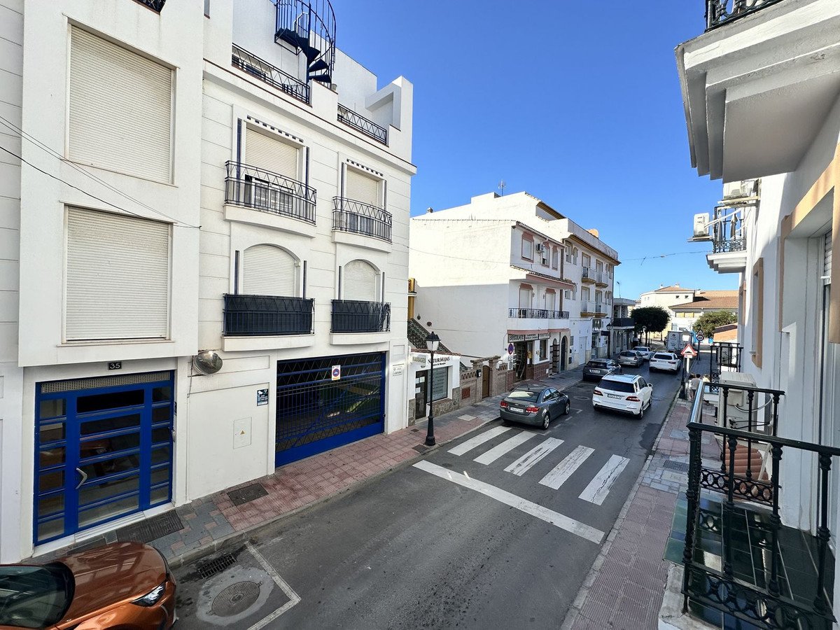 View down a street with parked cars and buildings on either side. Features balconies and street access doors.
