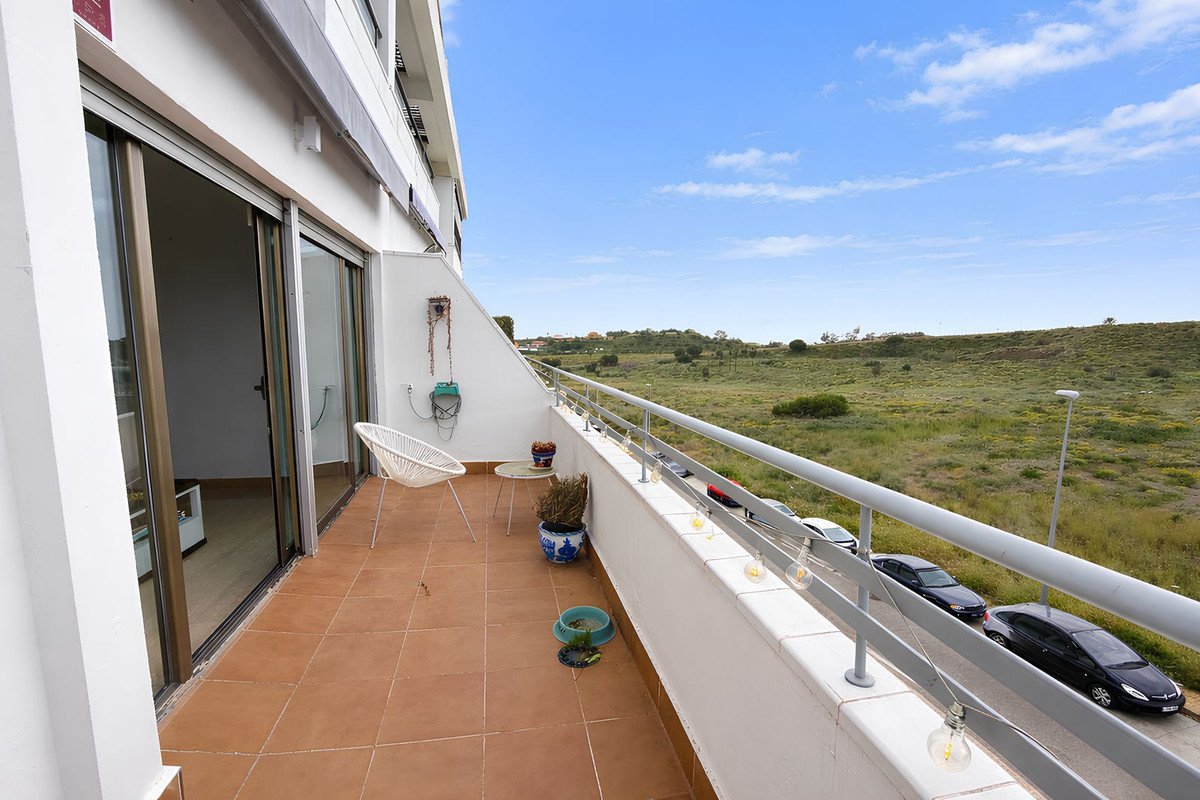 Balcony with tiled floor, white chair, and railing overlooking green hills and parked cars under a blue sky.