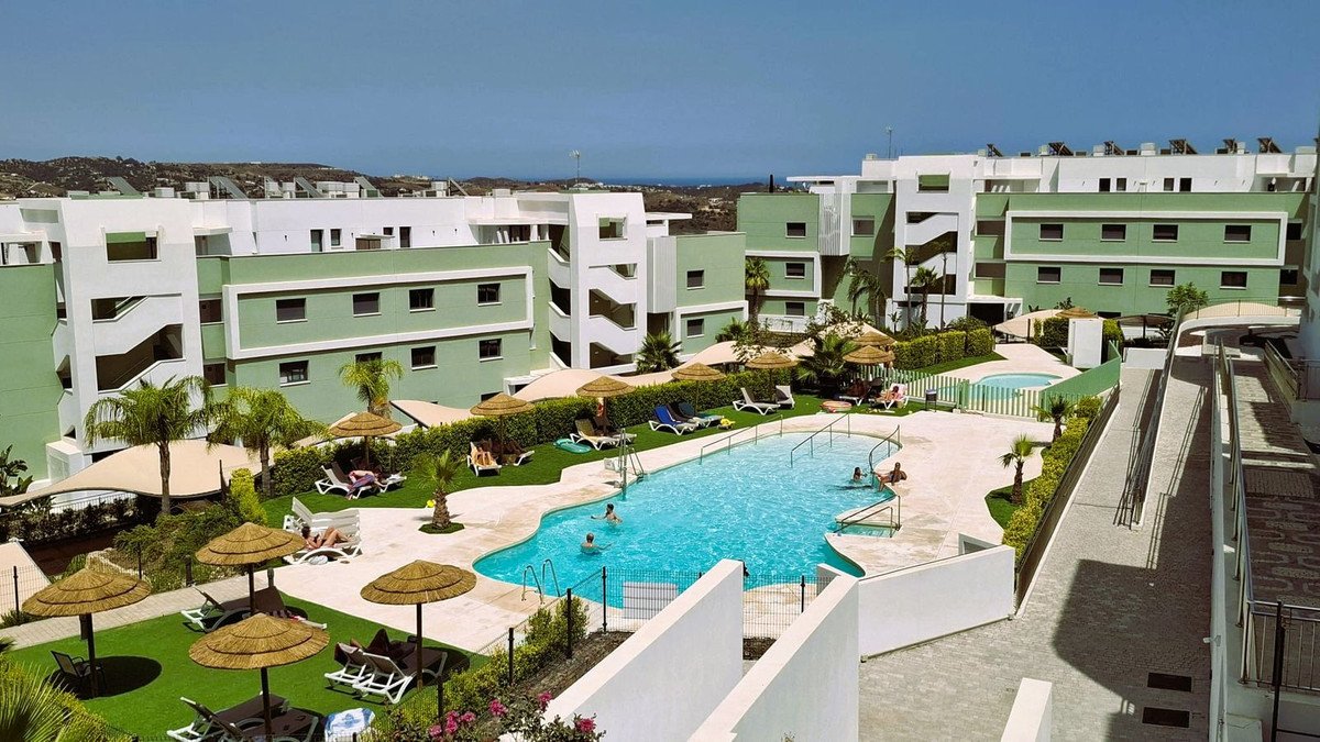 Apartment buildings overlook a resort-style swimming pool with lounge chairs and umbrellas on a sunny day.