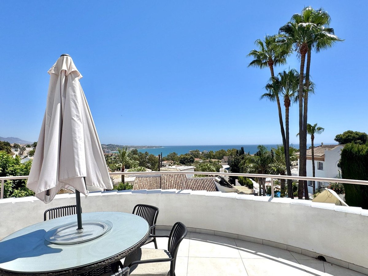 A balcony with a table and umbrella overlooks a sea view with palm trees under a clear blue sky.