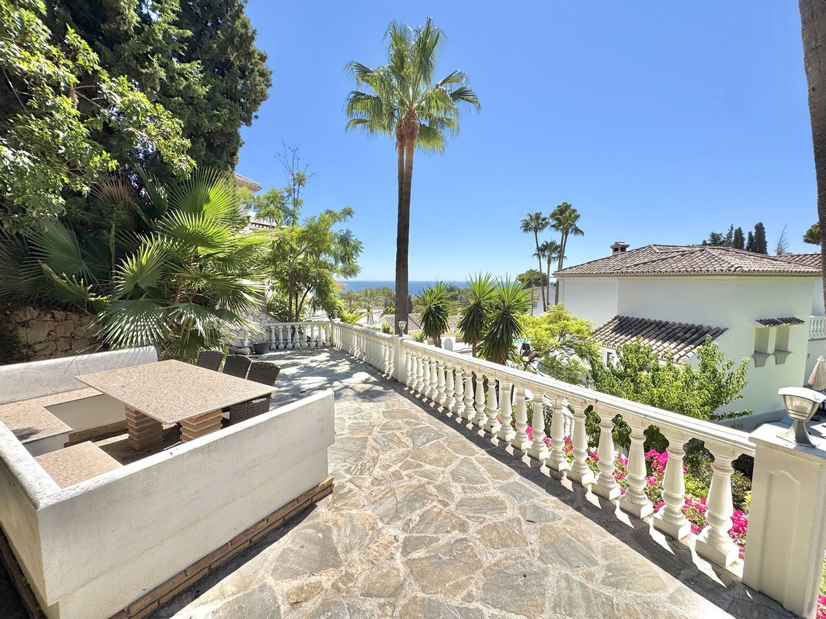 Terrace with built-in seating and dining table, overlooking the sea with palm trees and white villa.