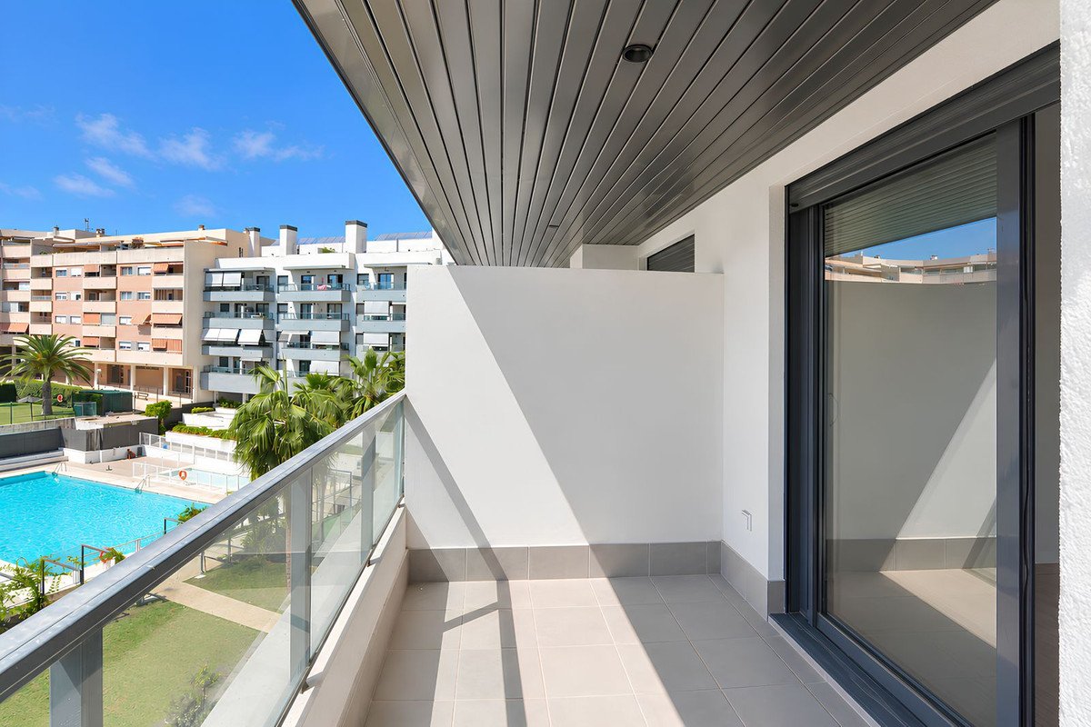 Modern balcony with glass railings overlooking a swimming pool and apartment buildings under a clear blue sky.