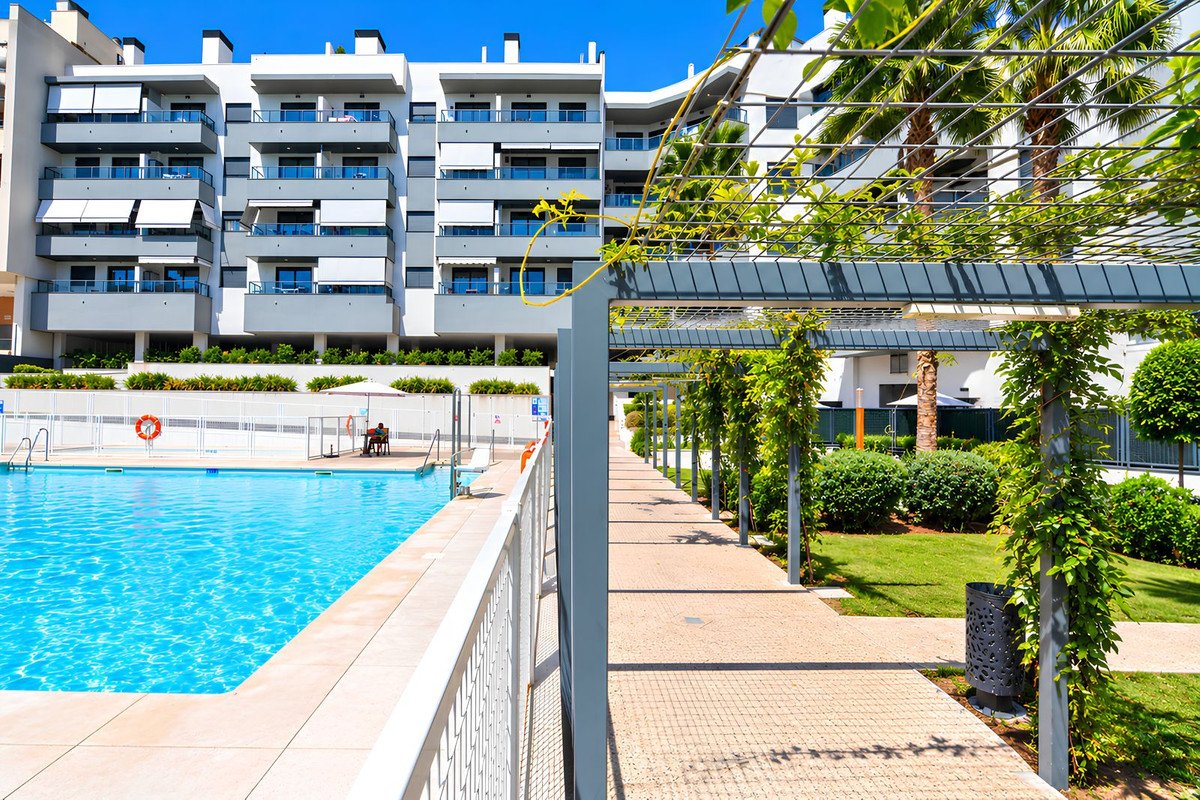 Modern apartment building with balconies overlooking a bright blue swimming pool and a landscaped garden path with pergolas and lush with greenery.