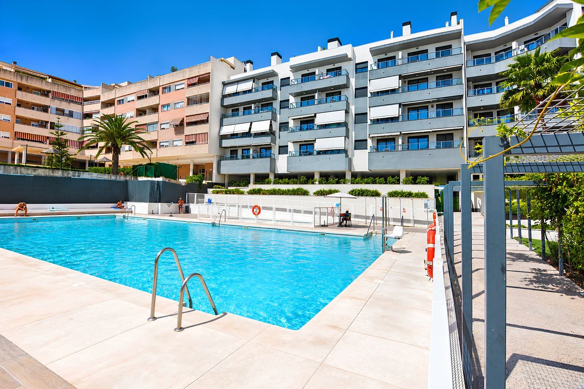 Outdoor swimming pool area with lounge chairs, modern apartment buildings, and lush greenery under a clear blue sky.