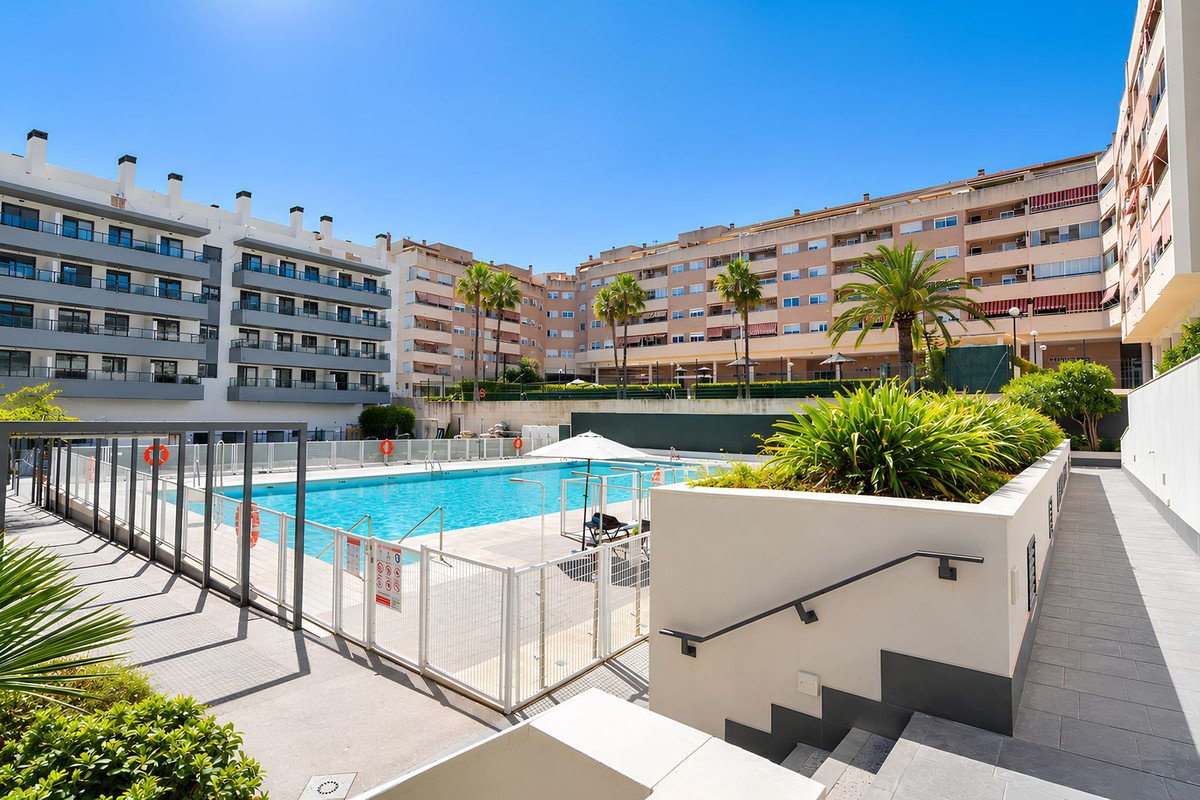 Large communal swimming pool surrounded by apartment buildings and palm trees under a clear blue sky.