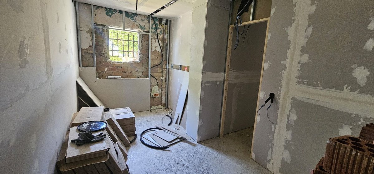 Interior room undergoing renovation with exposed brick, drywall, and construction materials visible. Window shows greenery outside.