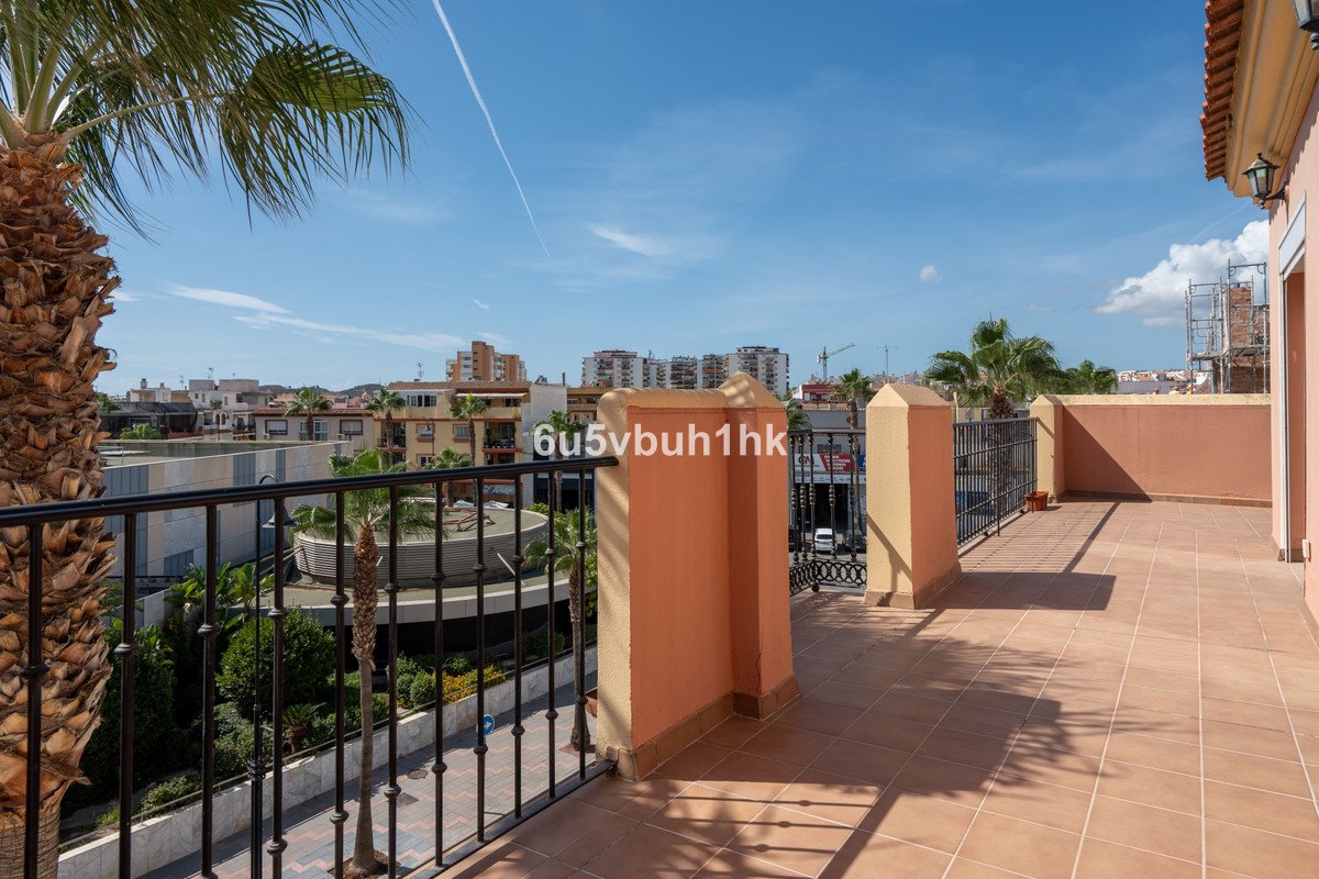 Spacious tiled terrace with black metal railings overlooking a street and buildings, featuring a palm tree and clear blue sky.