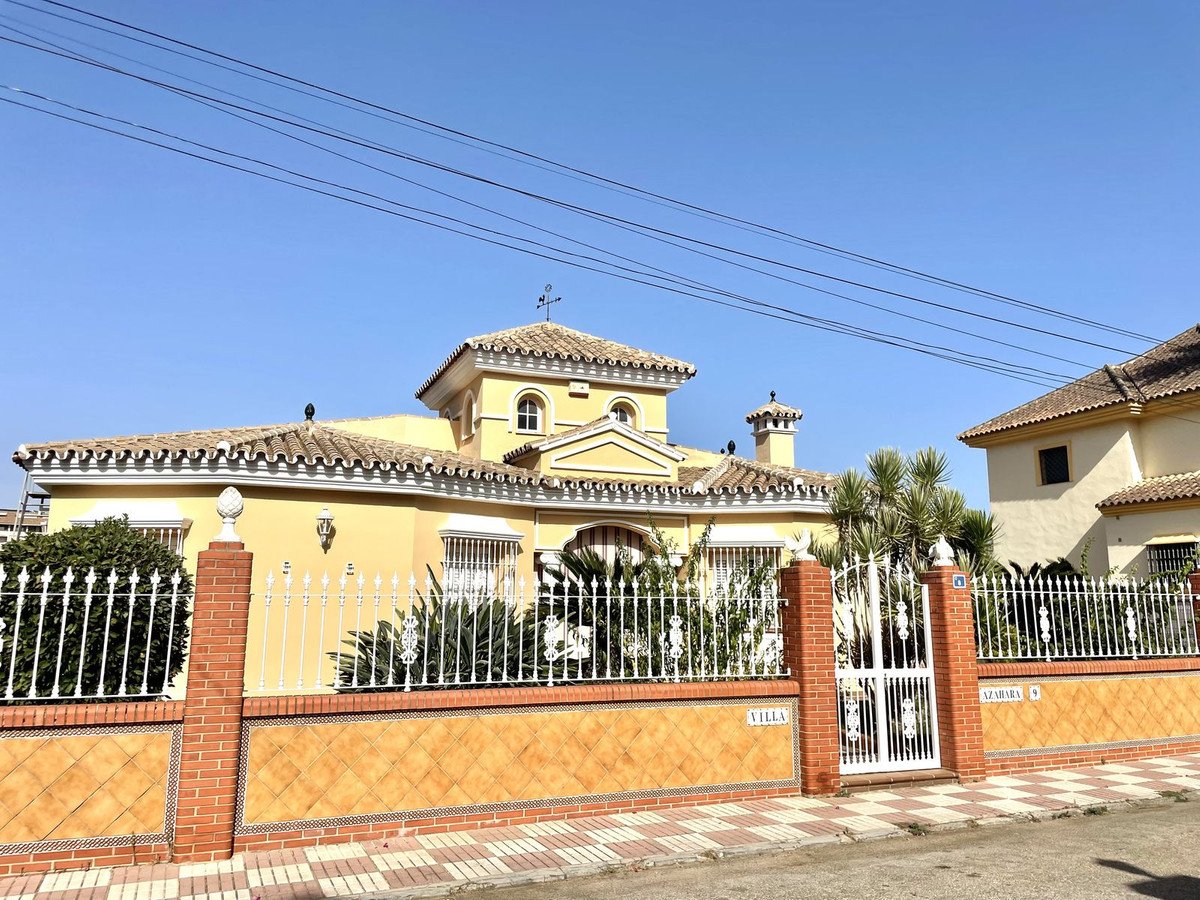Exterior view of a two-story yellow villa with a white fence, ornate gate, and palm trees under a clear blue sky.