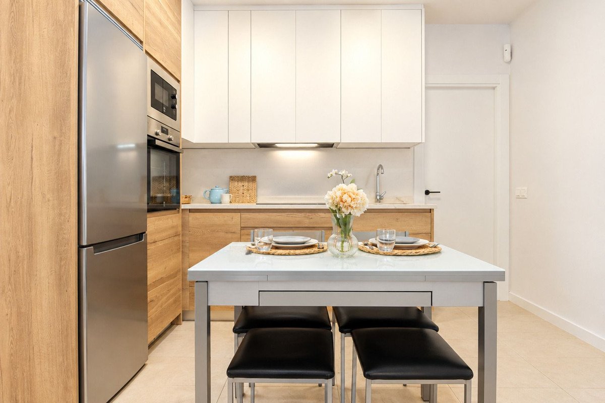 A modern kitchen with a dining table set for two, featuring sleek cabinetry, a stainless steel refrigerator, and a sink.