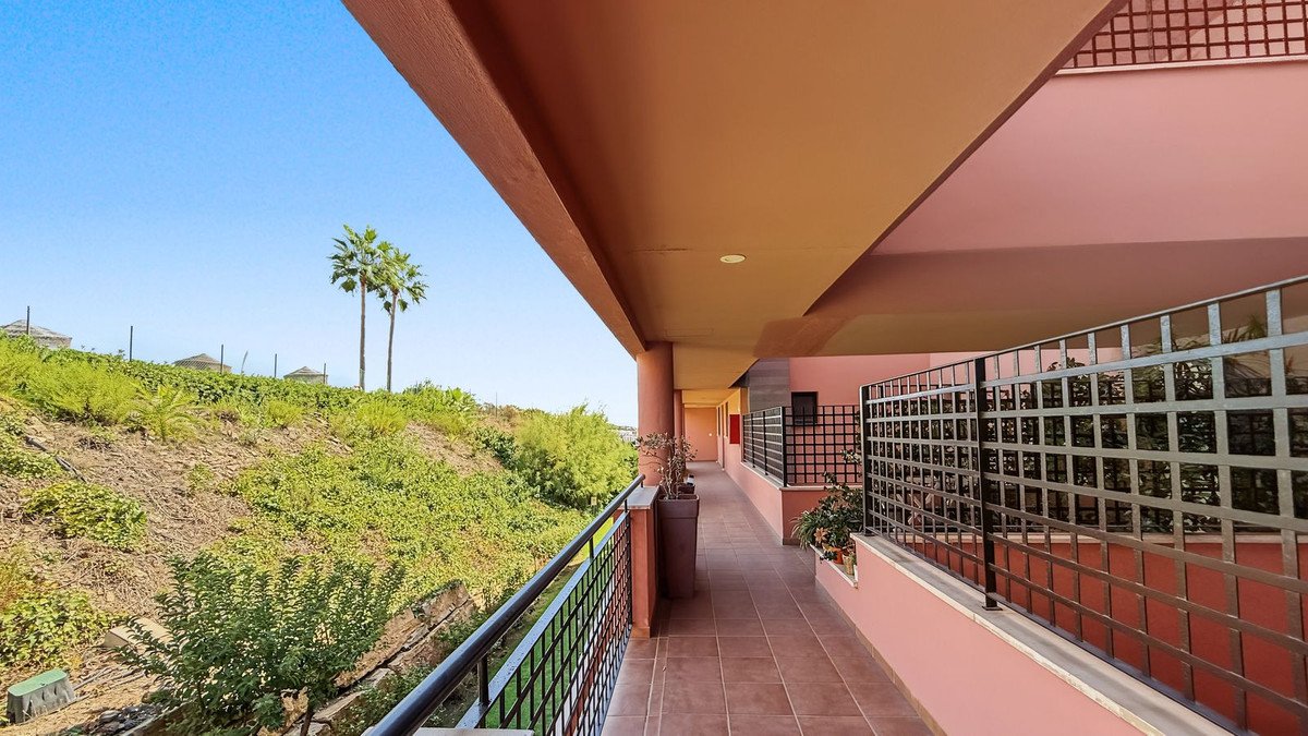 A long, pink-walled balcony with a metal railing overlooks a green hillside with palm trees under a clear blue sky.